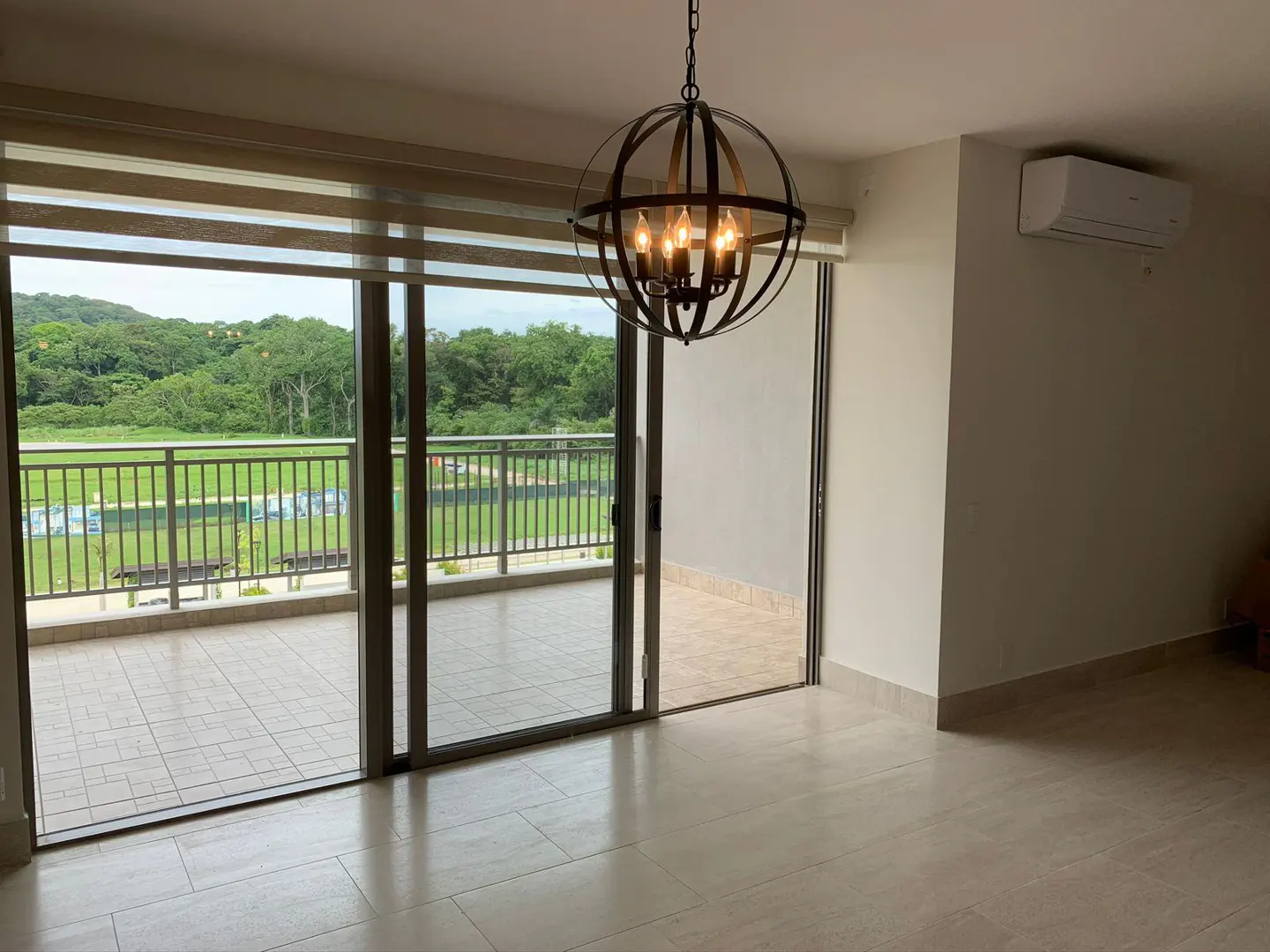 Bright, empty room with tile floor, sliding glass doors to balcony with green view. Black orb chandelier hangs from ceiling.