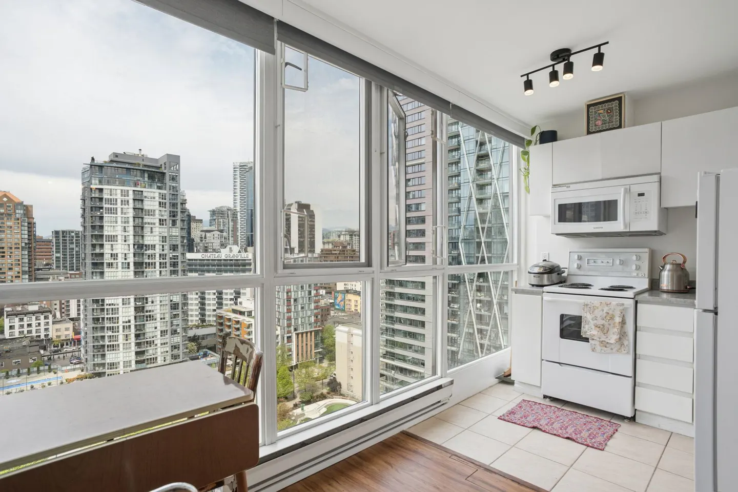 A bright, modern kitchen with white cabinets, appliances, and a large window overlooking a cityscape. A wooden table sits near the window.