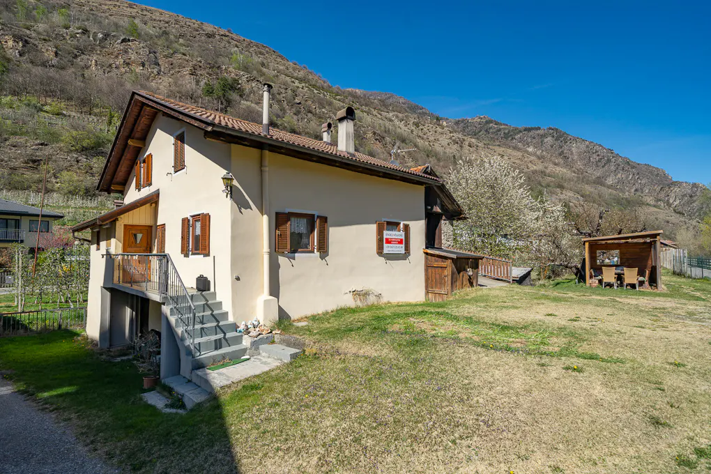 Exterior view of a cream-colored house with brown shutters, a "For Sale" sign, and a mountain backdrop.