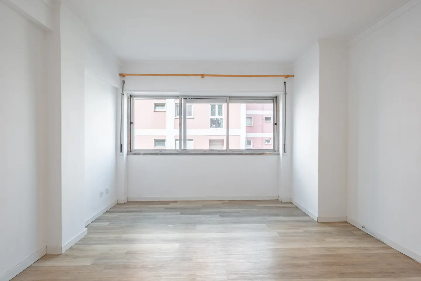 Empty room with white walls and light wood floor. A window shows a building outside.