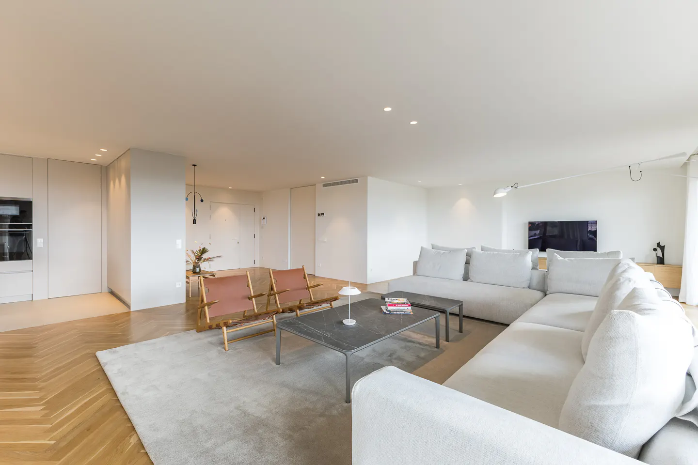 Bright, modern living room with white walls, herringbone wood floors, and a large white sectional sofa. Two leather chairs face a gray coffee table.