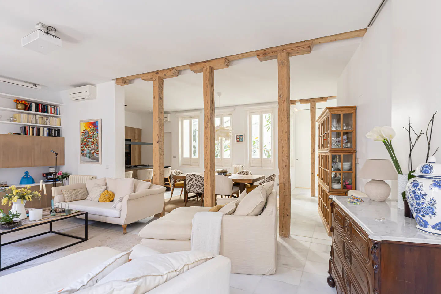 Bright, open-plan living and dining area with white sofas, wooden beams, and marble floors. A wooden cabinet and dining table are visible.