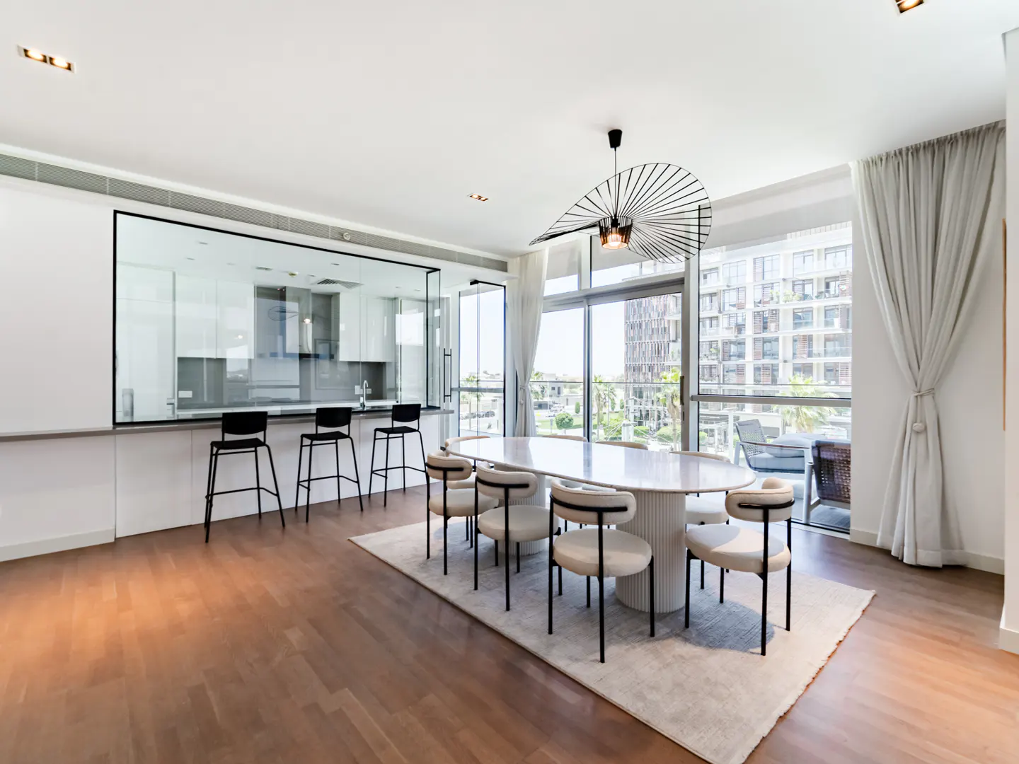 Bright, modern dining area with an oval table, chairs, and a unique light fixture. A kitchen is visible through a glass partition.