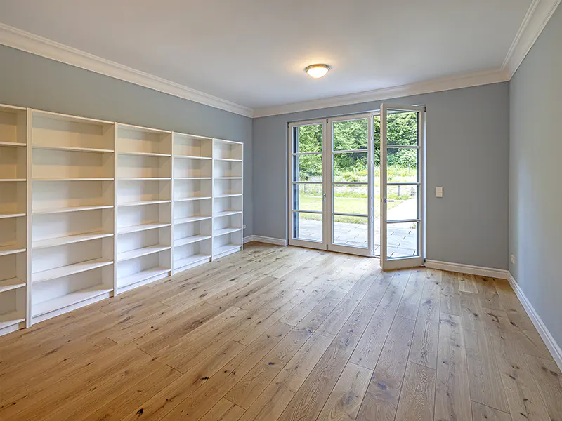 Bright room with wood floors, light blue walls, and white trim. White bookshelves line one wall, and glass doors open to a green yard.