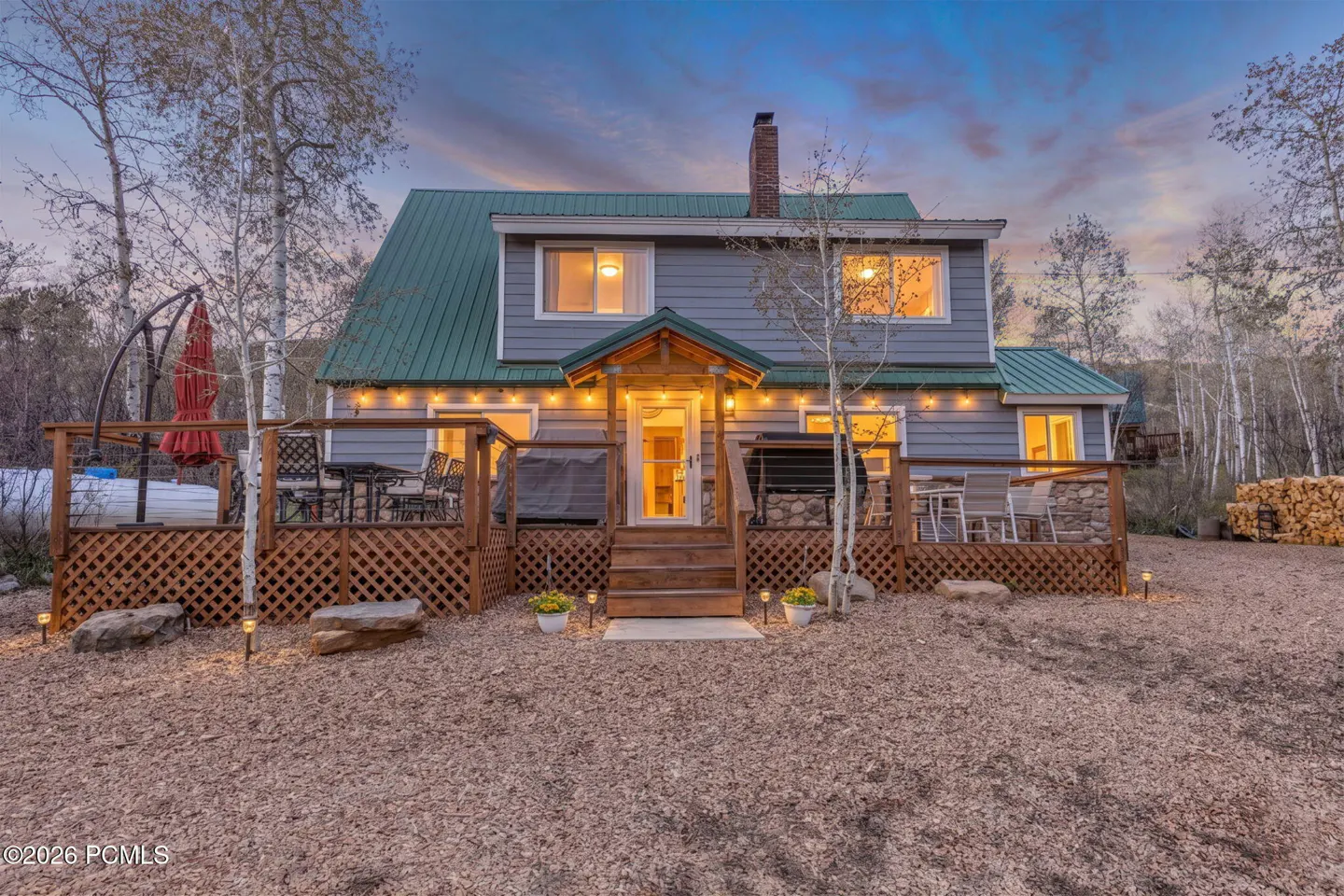 Exterior view of a two-story gray house with a green roof and a wooden deck at dusk. String lights illuminate the deck.