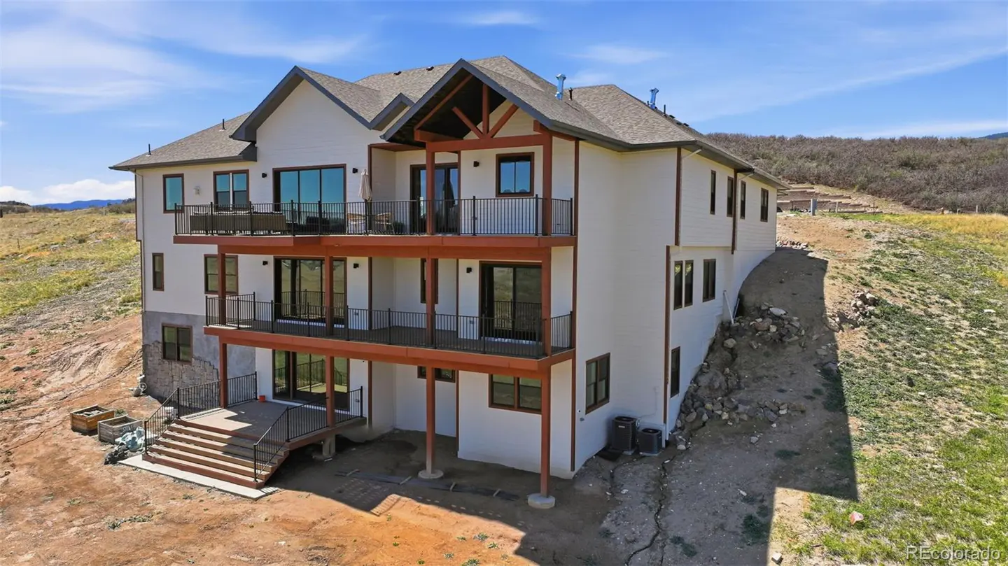 Rear view of a three-story white house with brown trim and black railings on multiple decks. The house is on a hillside with a blue sky.