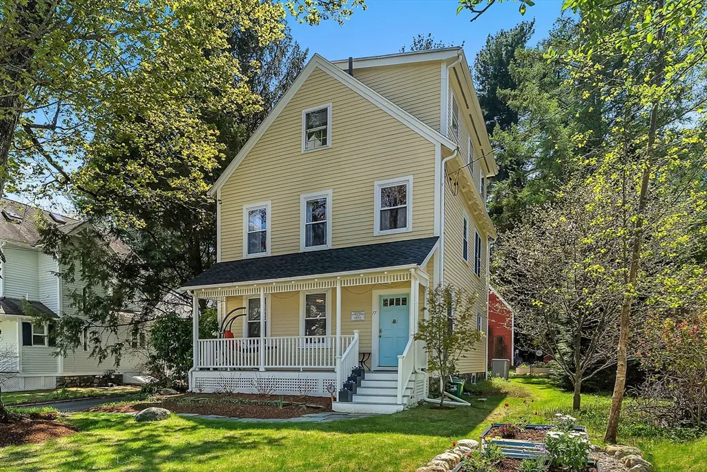 Two-story yellow house with a white porch and a light blue front door, surrounded by green trees and lawn.