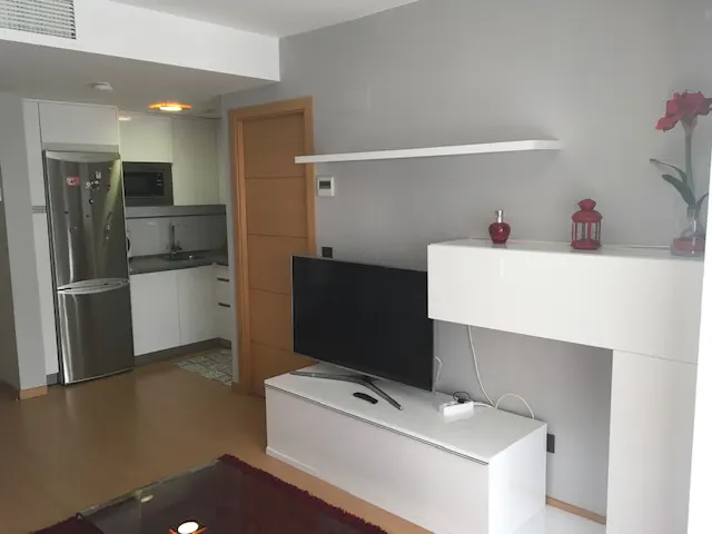 Living room with a TV, white cabinets, and a stainless steel refrigerator. A red flower and lantern sit on the cabinet.