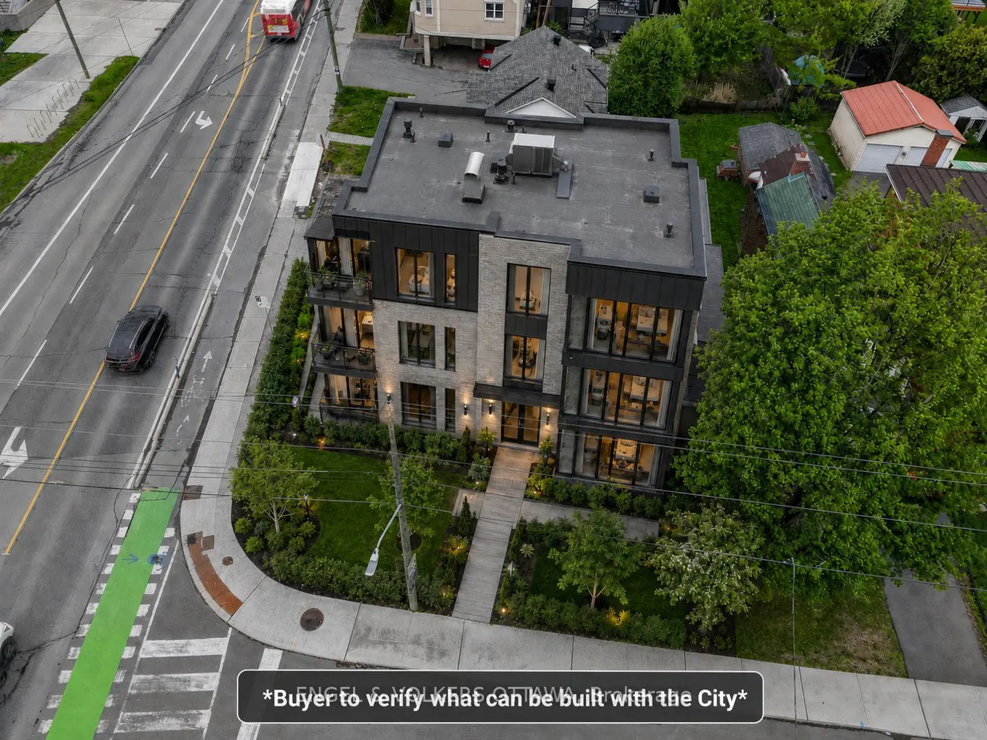 Aerial view of a modern, three-story apartment building with large windows and stone accents, surrounded by greenery and a city street.