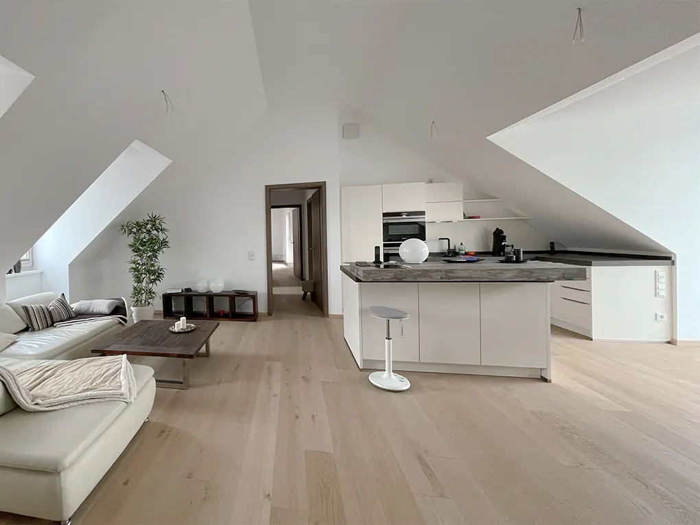 Open-concept living space with white walls, light wood floors, and sloped ceilings. A white sofa, kitchen island, and doorway are visible.