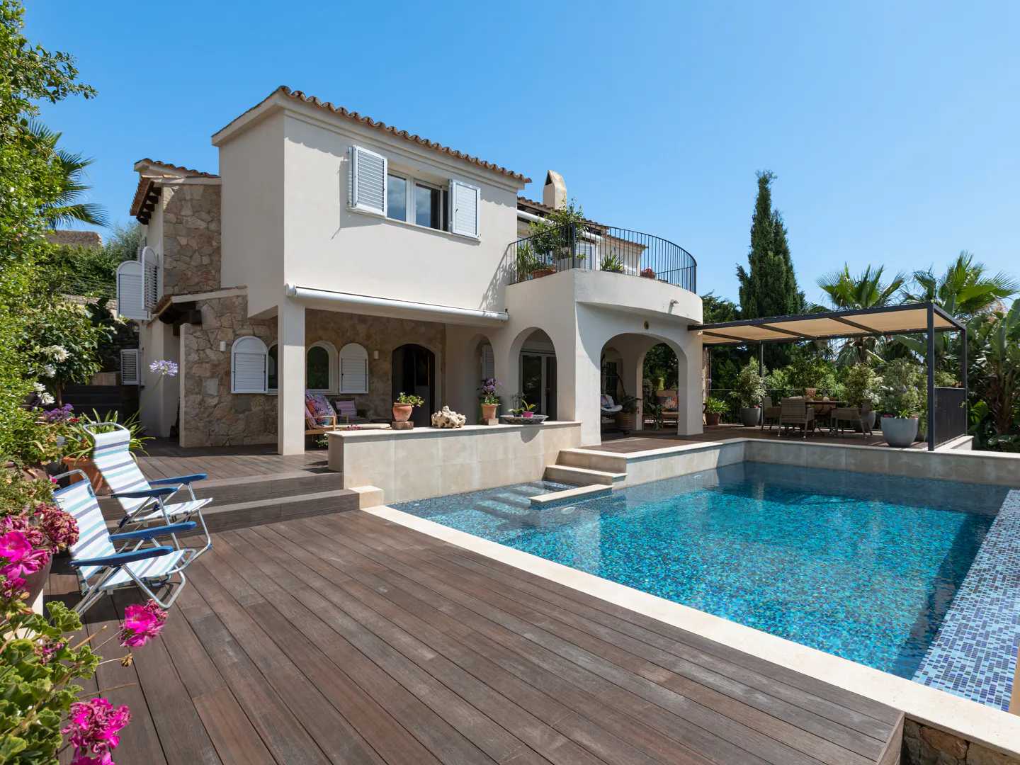 Luxury home with a pool. The house is two stories with a balcony and a stone facade. The pool is surrounded by a wooden deck and lounge chairs.