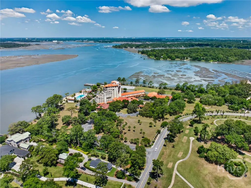 Aerial view of a waterfront resort with a red tile roof, surrounded by green trees and a golf course, under a blue sky.