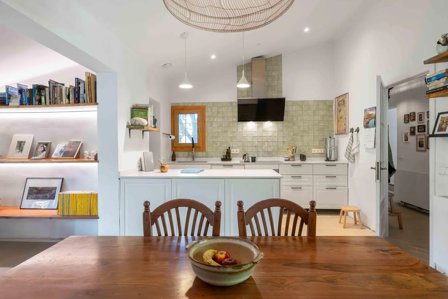 A bright, modern kitchen with a wooden table and chairs in the foreground. The kitchen features white cabinets, a tiled backsplash, and open shelving.