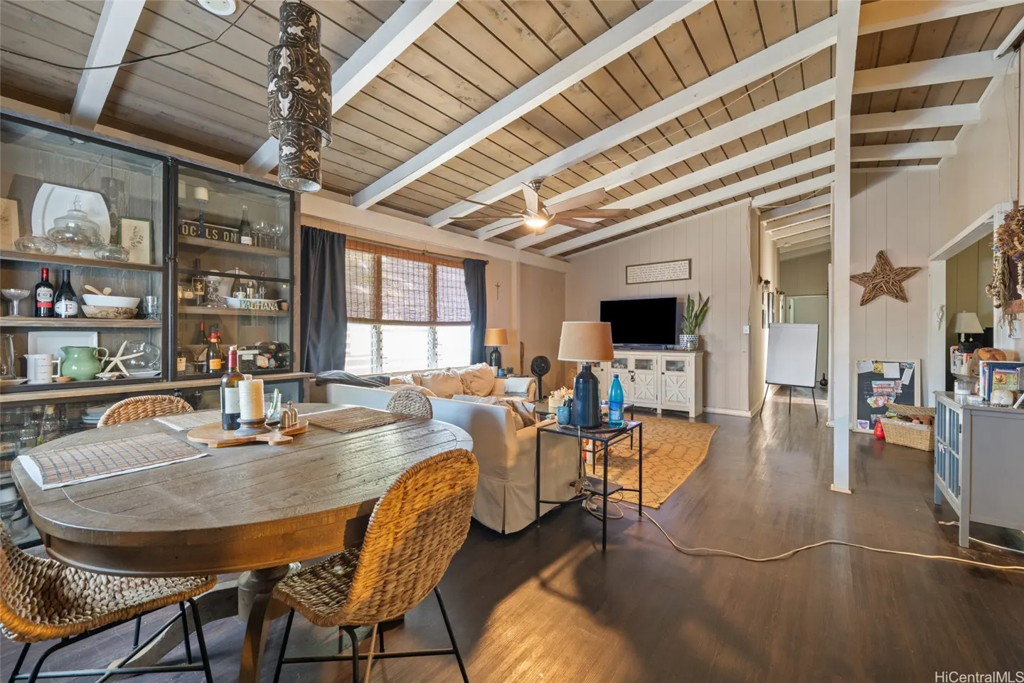 Living room with a wood table, wicker chairs, and a glass cabinet filled with bottles and dishes. The ceiling is wood with white beams.