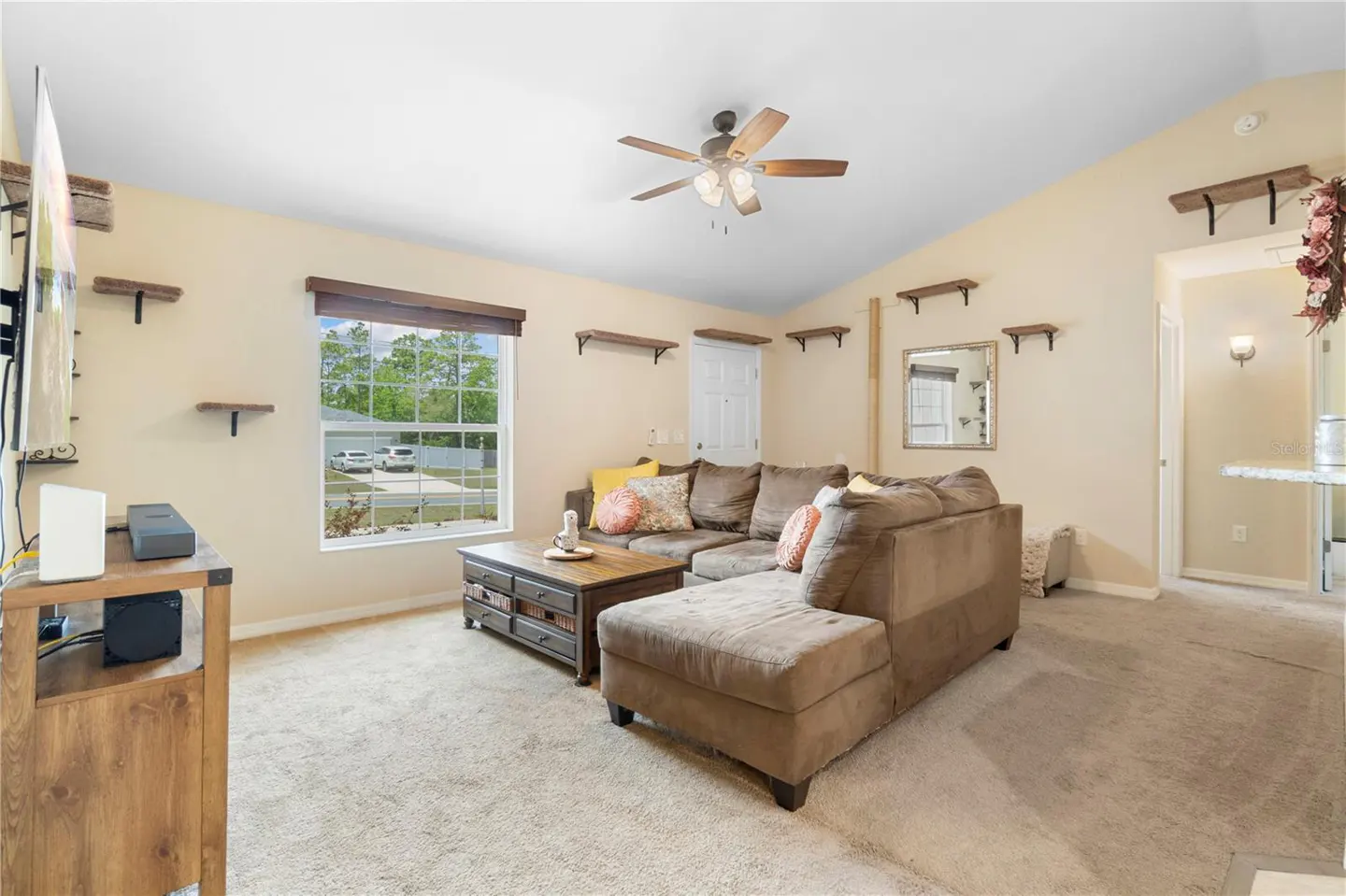 Living room with beige walls, carpet, and a brown sectional sofa. A wooden coffee table sits in front of the sofa, and a window provides natural light.