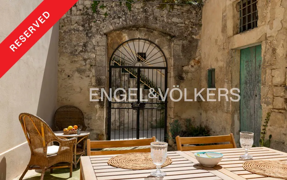 A rustic stone courtyard with a wooden dining table, wicker chairs, and an arched iron gate, marked as reserved.