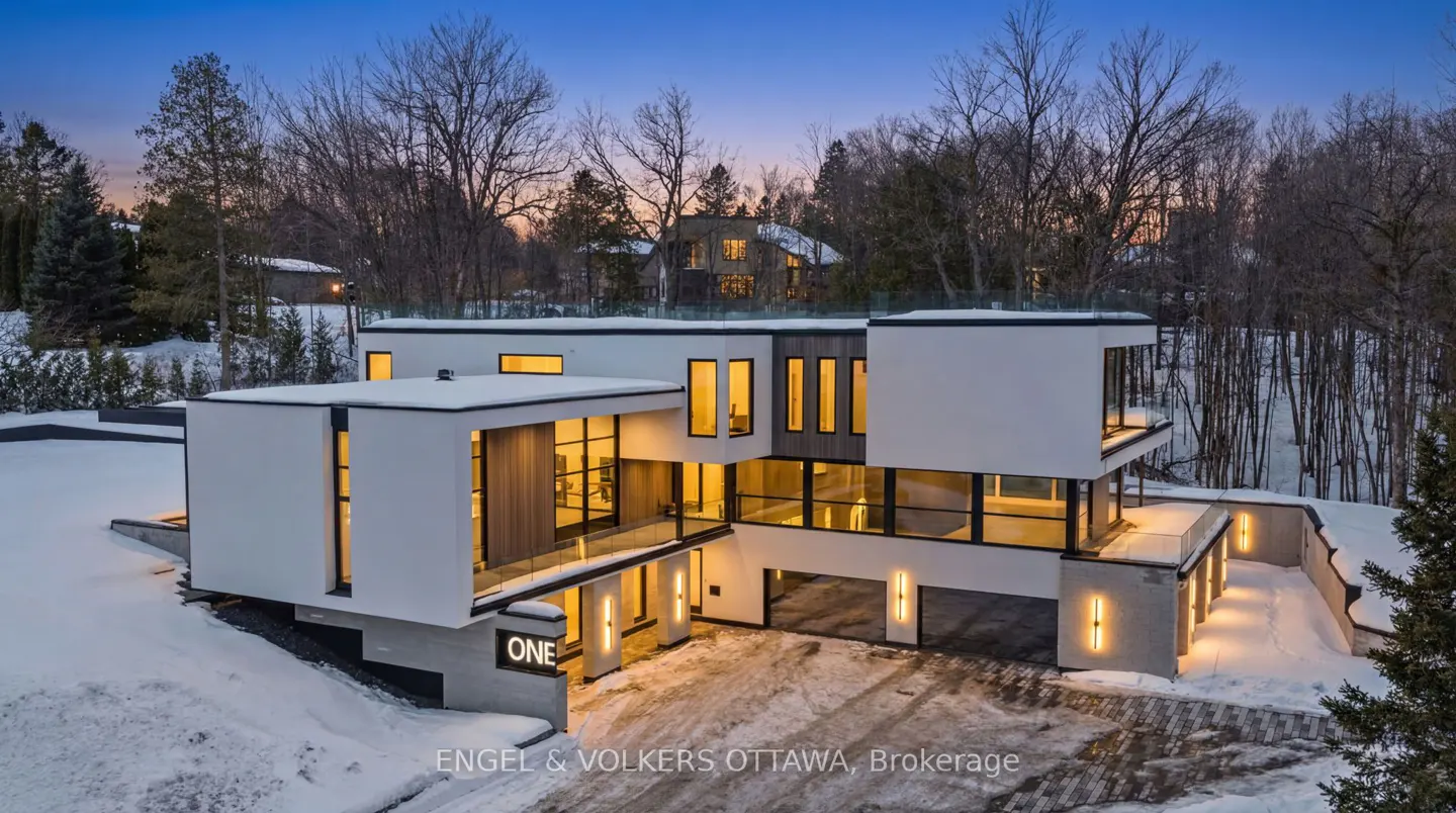 Modern white house with black framed windows, and a driveway covered in snow. Trees in the background.