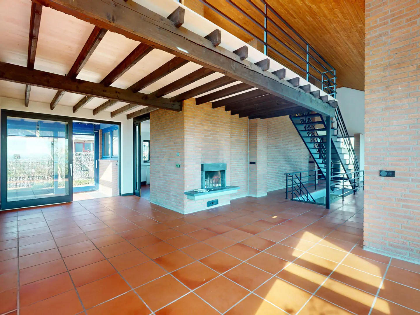 A spacious living room with brick walls, terracotta tile floors, and a wood-beamed ceiling. A modern black staircase leads to a loft area. A fireplace is centered on one wall. Sliding glass doors open to the outside.