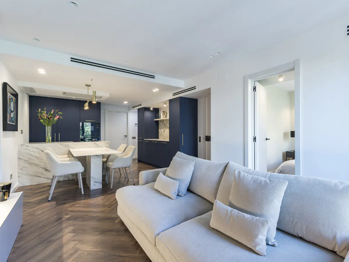 Open-concept living space with a gray sofa, wood floors, and a marble kitchen island with blue cabinets. A bedroom is visible through an open doorway.
