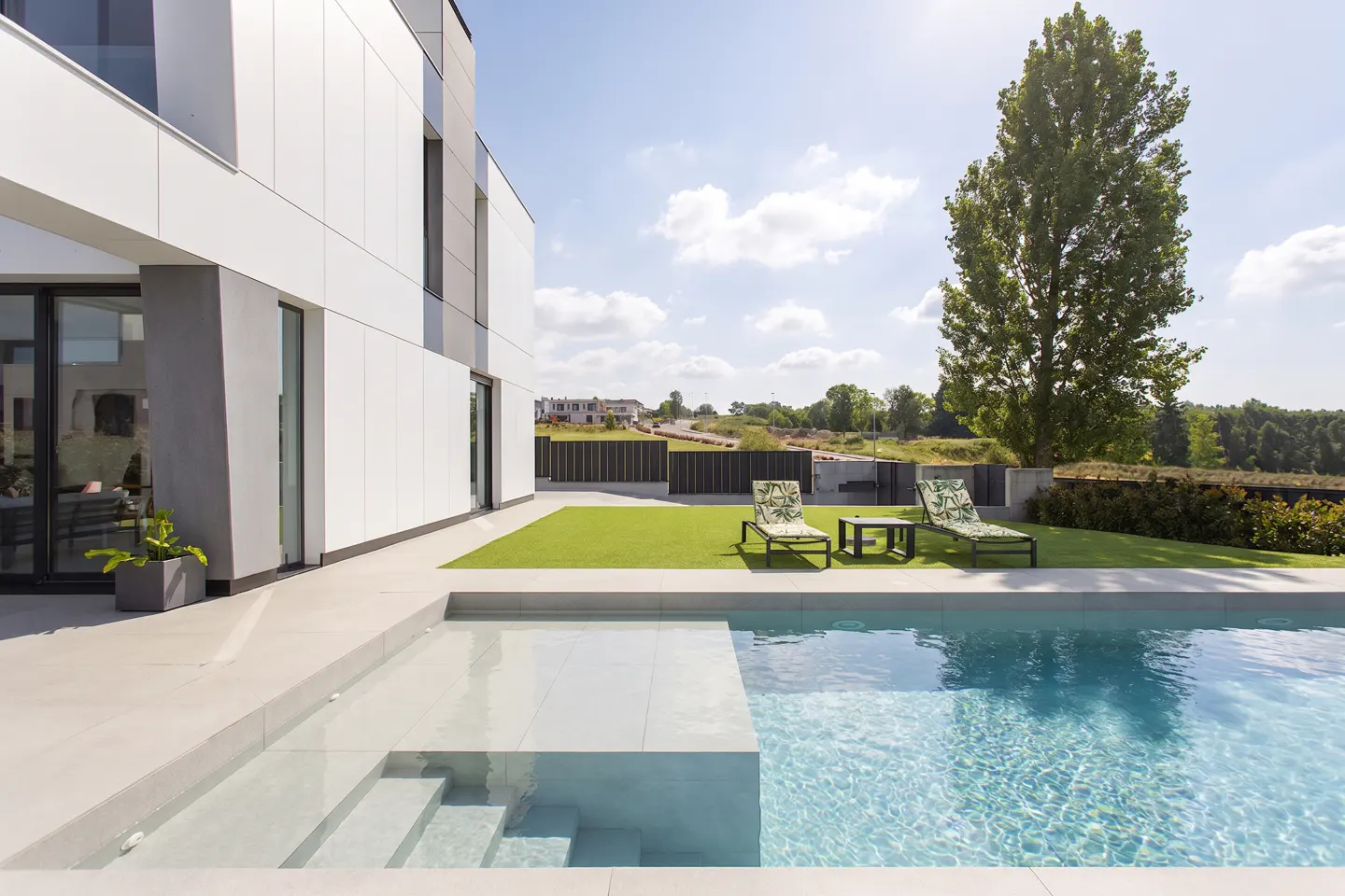 Modern home exterior with a pool, lounge chairs, and green lawn on a sunny day.