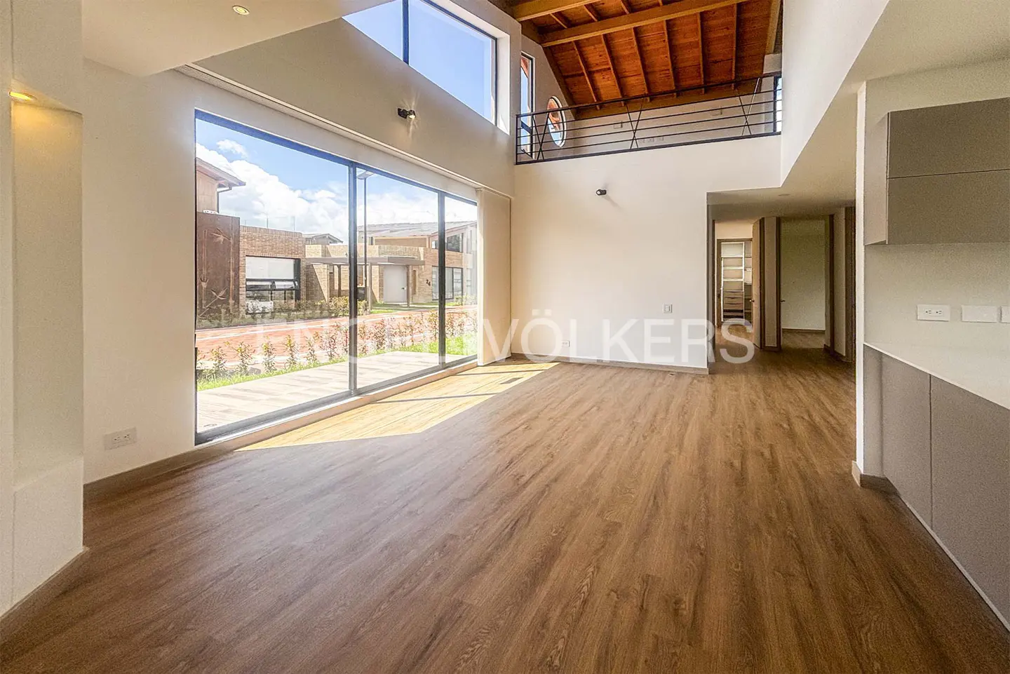 Bright, modern living room with wood floors, large windows, and a wooden ceiling. Balcony with black railing overlooks the space.