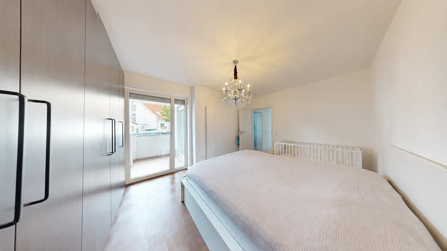Bedroom with white walls, wood floor, large bed, and balcony. A crystal chandelier hangs from the ceiling. A crib is in the corner.