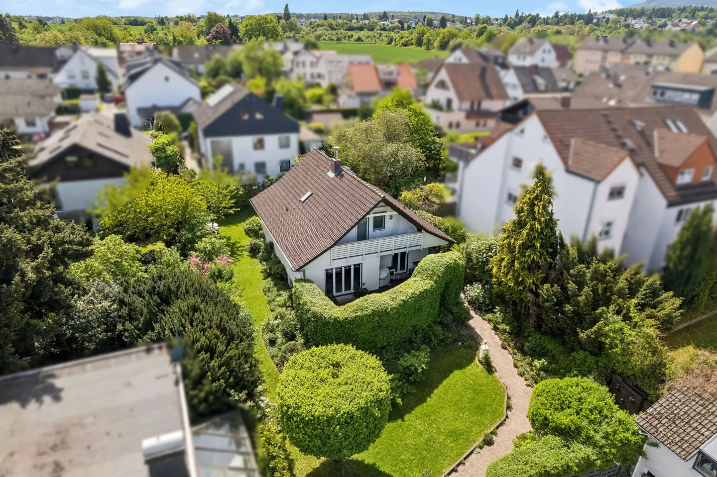 Aerial view of a white house with a brown roof, surrounded by lush green landscaping and other houses in the background.