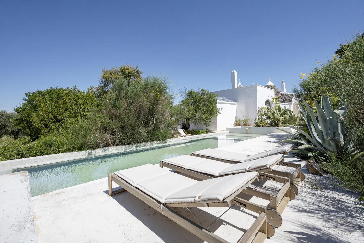Outdoor view of a white trullo house with a pool and lounge chairs under a clear blue sky.