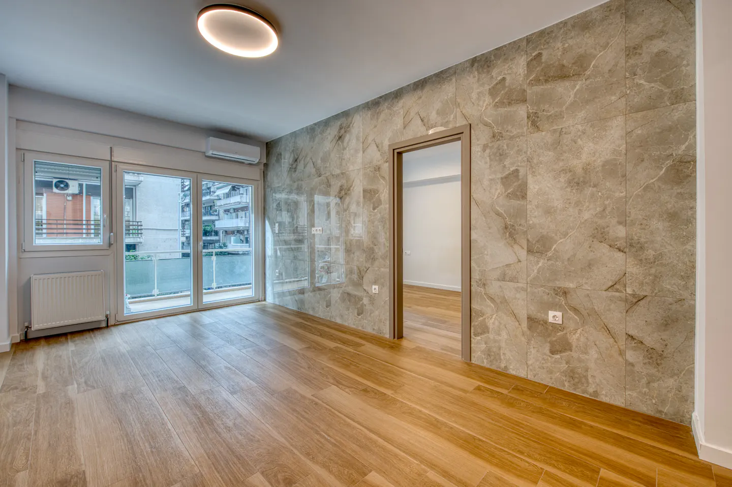 Bright, empty room with wood floors, marble accent wall, and sliding glass doors to a balcony overlooking a canal.