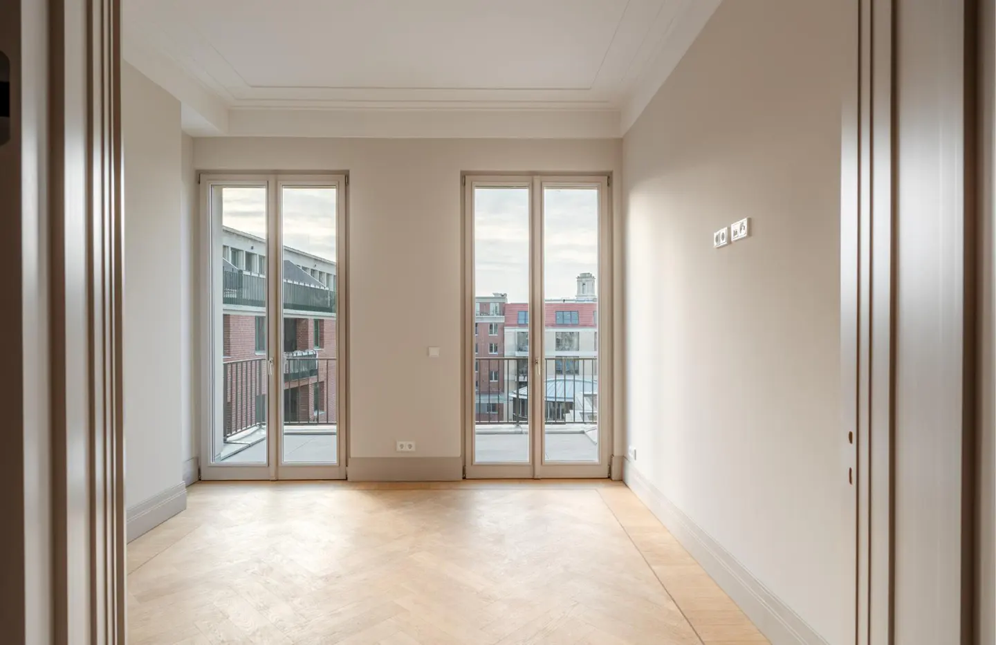 Empty room with light beige walls, herringbone wood floors, and two glass doors opening to a balcony with city views.