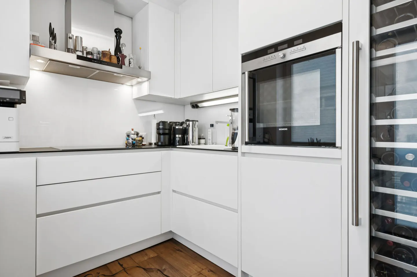 A modern, white kitchen with stainless steel appliances, including an oven and wine cooler. The floor is made of dark wood.