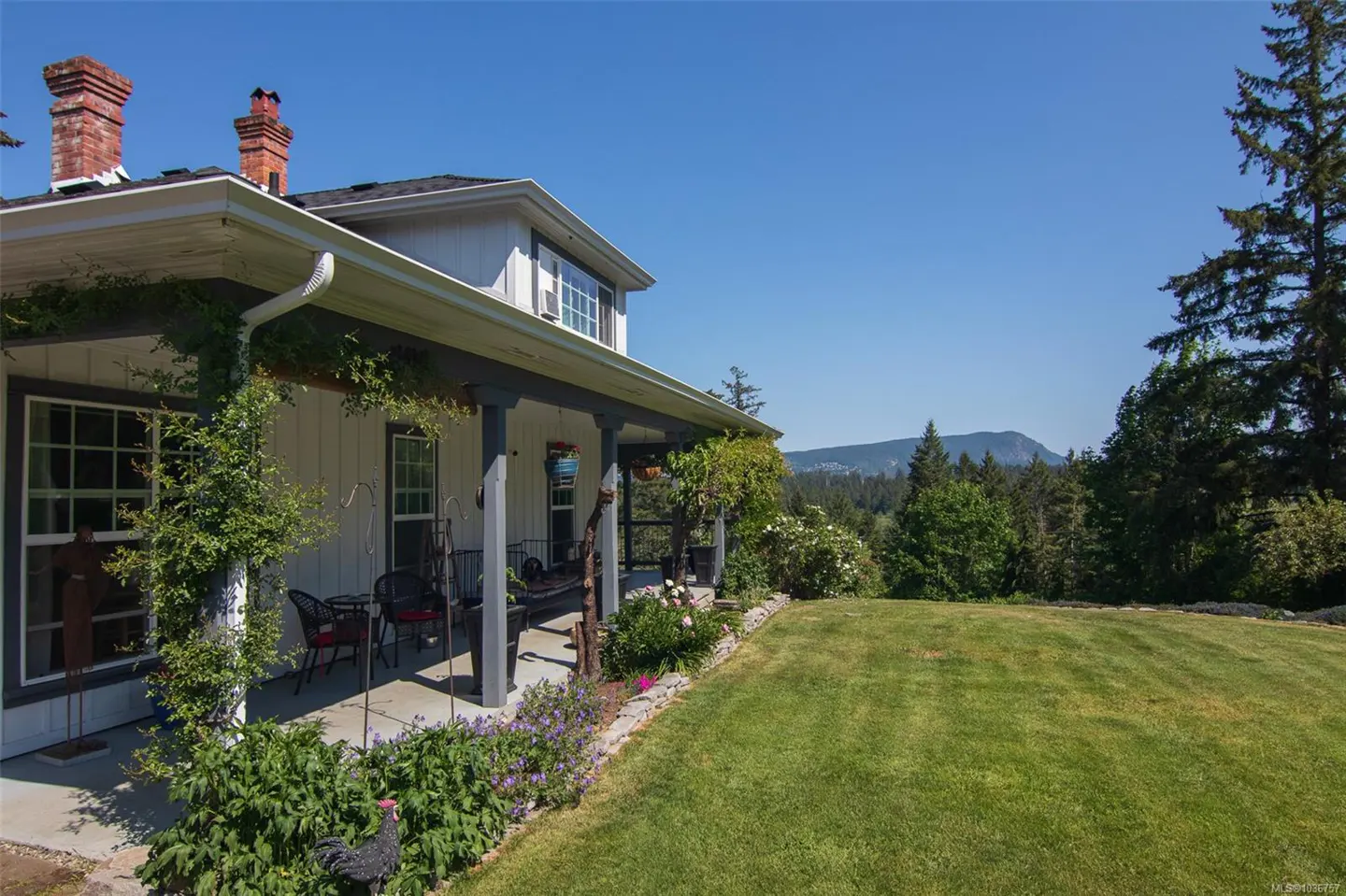Exterior view of a white farmhouse with a covered porch, green lawn, and mountain views under a clear blue sky.