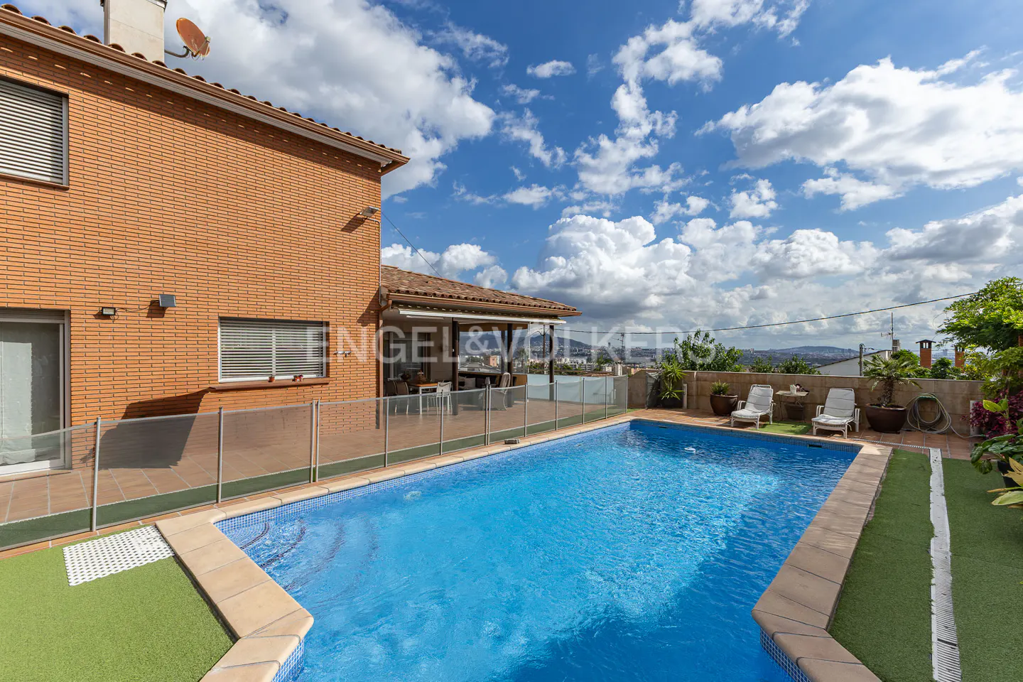 Backyard view of a brick house with a blue tiled pool, patio furniture, and a clear blue sky with white clouds.