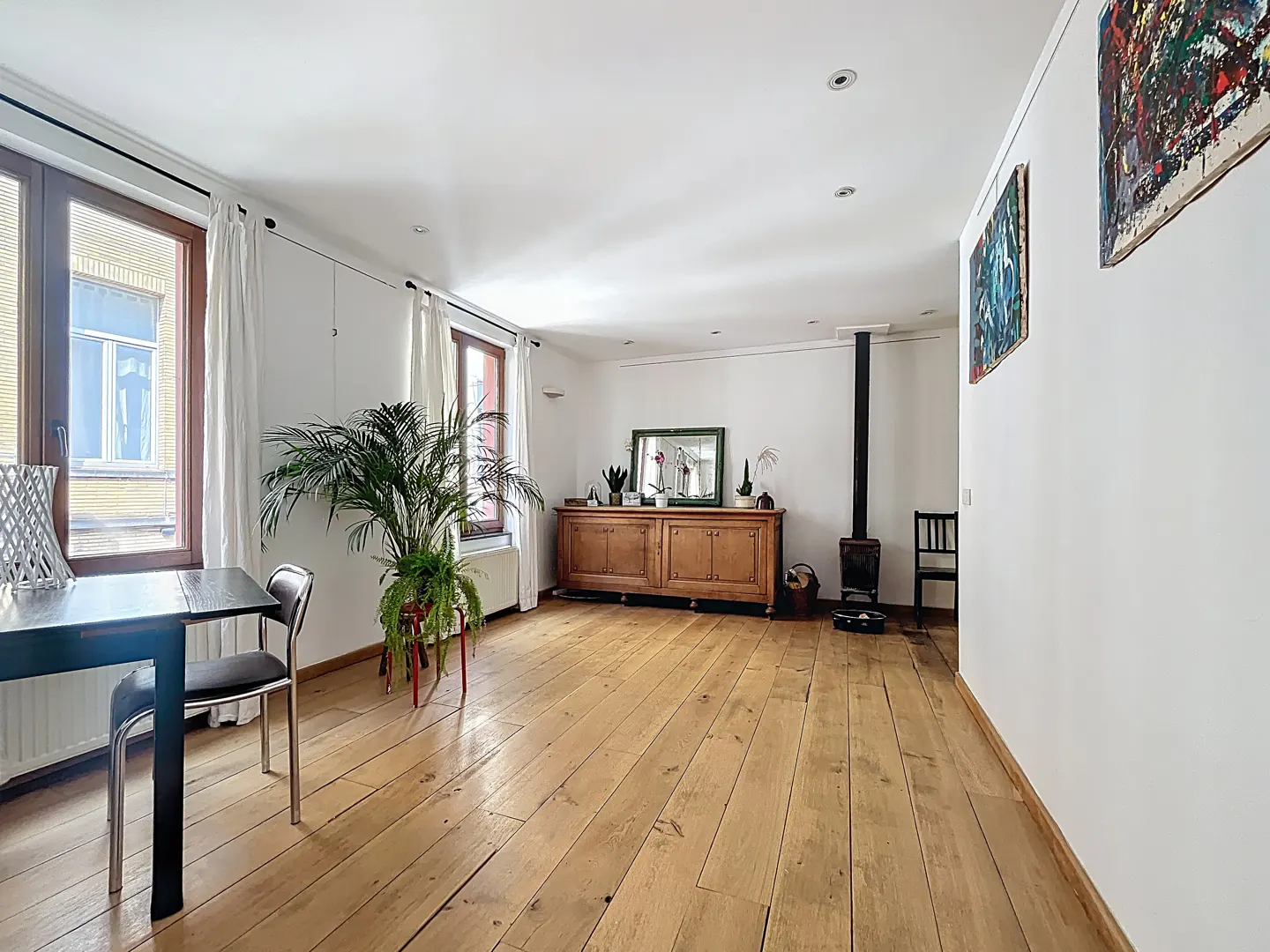 Bright living room with wood floors, white walls, and windows with white curtains. A table, chairs, cabinet, and artwork are visible.