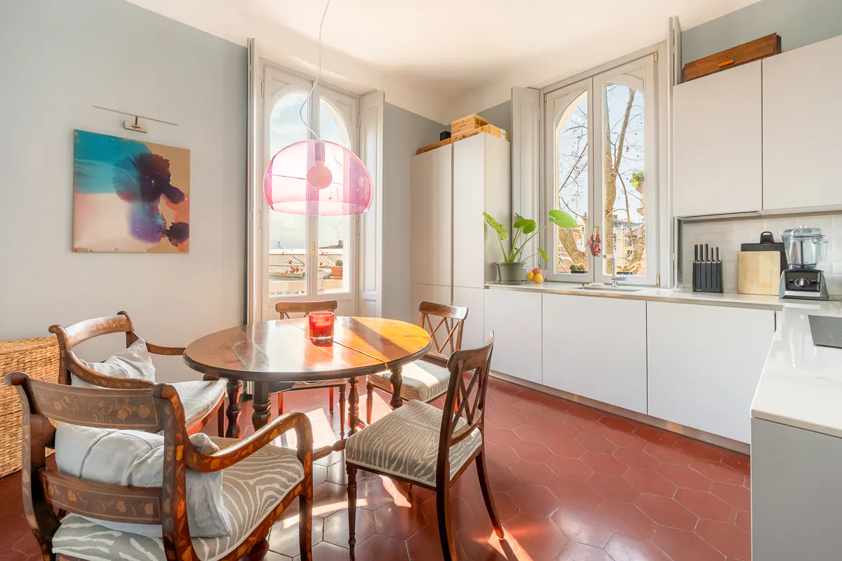 Bright kitchen with a round wooden table, chairs, and a pink pendant light. White cabinets line the wall, and sunlight streams through arched windows.