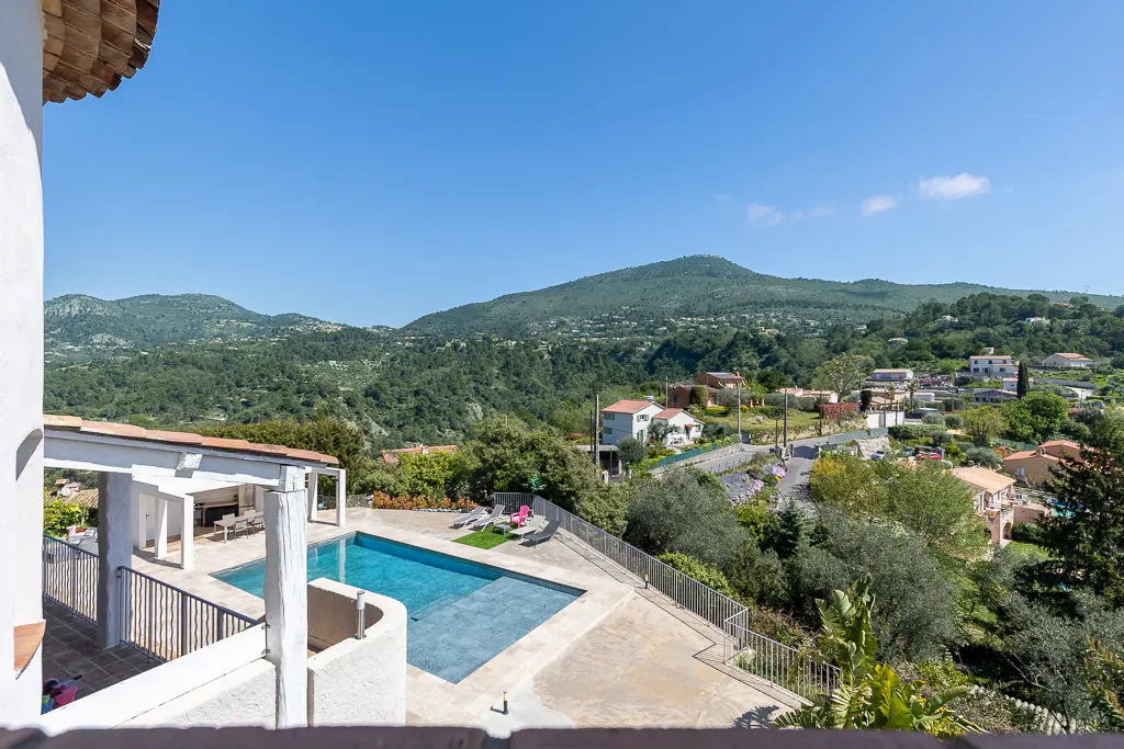 View from a terrace overlooking a blue swimming pool, lush green hills, and a small village under a clear blue sky.