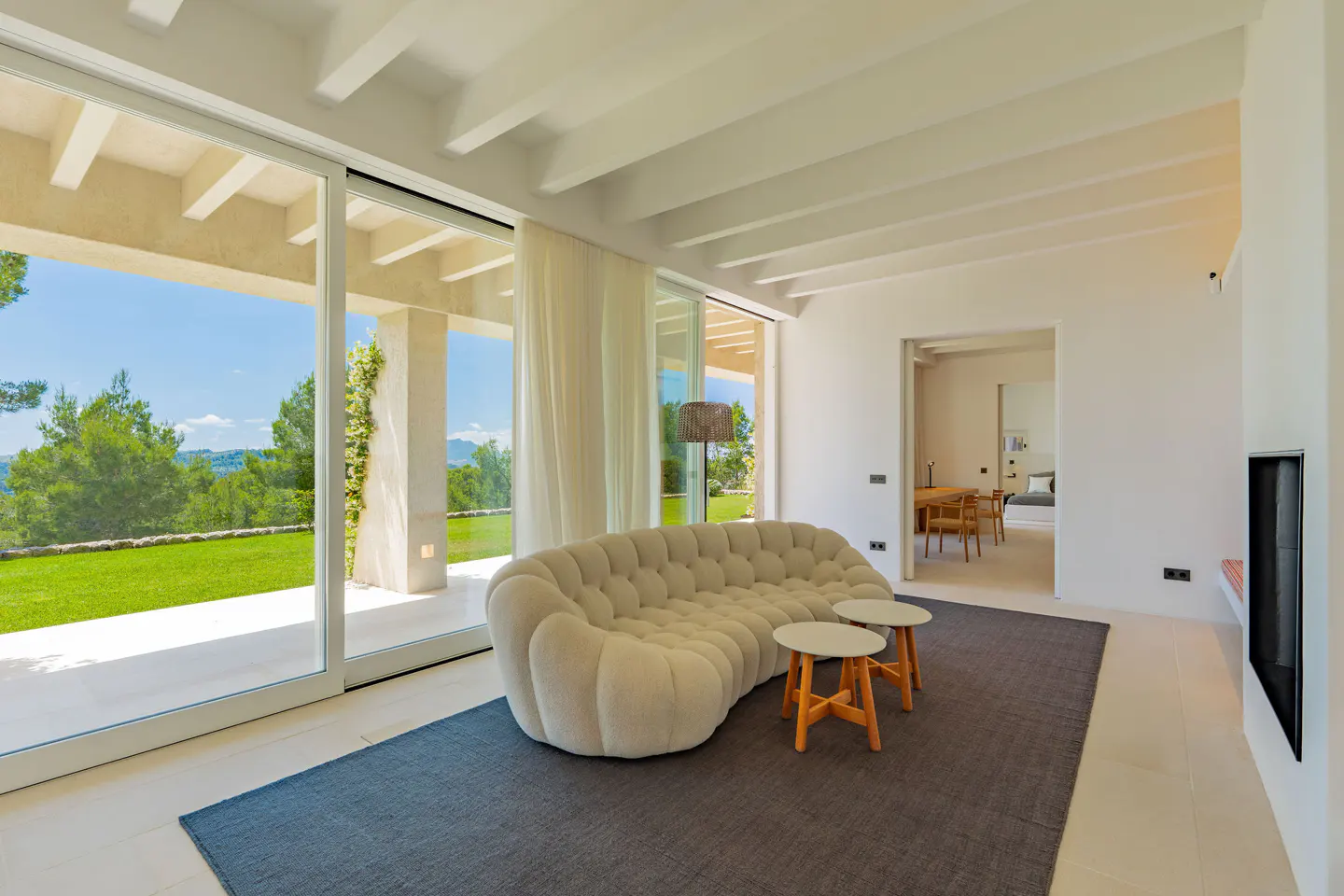 Bright living room with a curved, tufted sofa, two small tables, and a gray rug. Large sliding glass doors open to a green lawn and trees.
