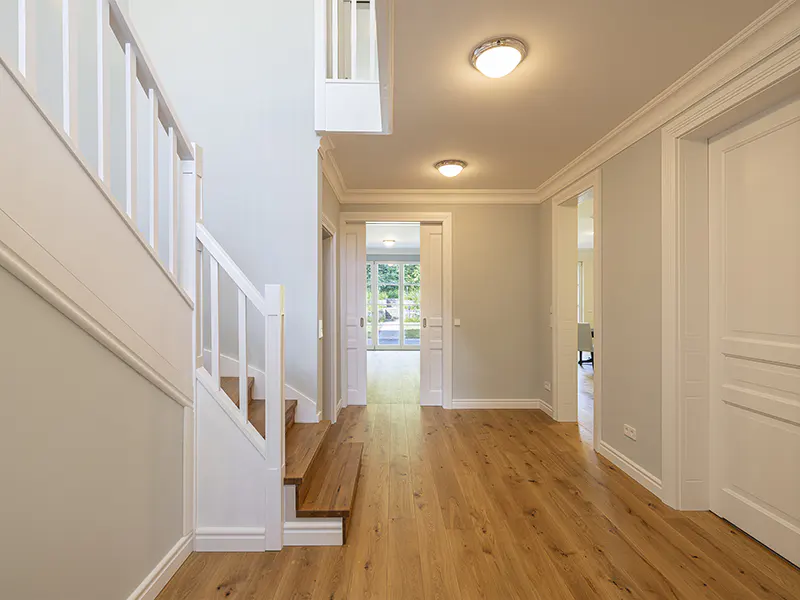 Bright hallway with wood floors, white trim, and pale gray walls. A staircase is on the left, and a doorway leads to a garden view.