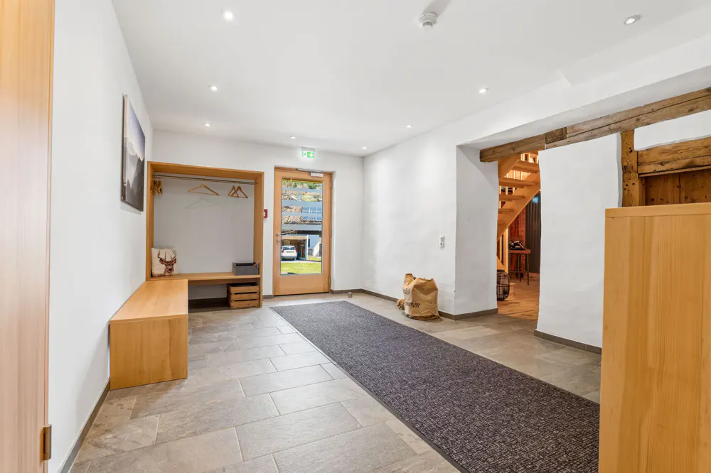 Entryway with light wood bench, coat rack, and gray rug leading to a glass door. A wooden staircase is visible in the background.