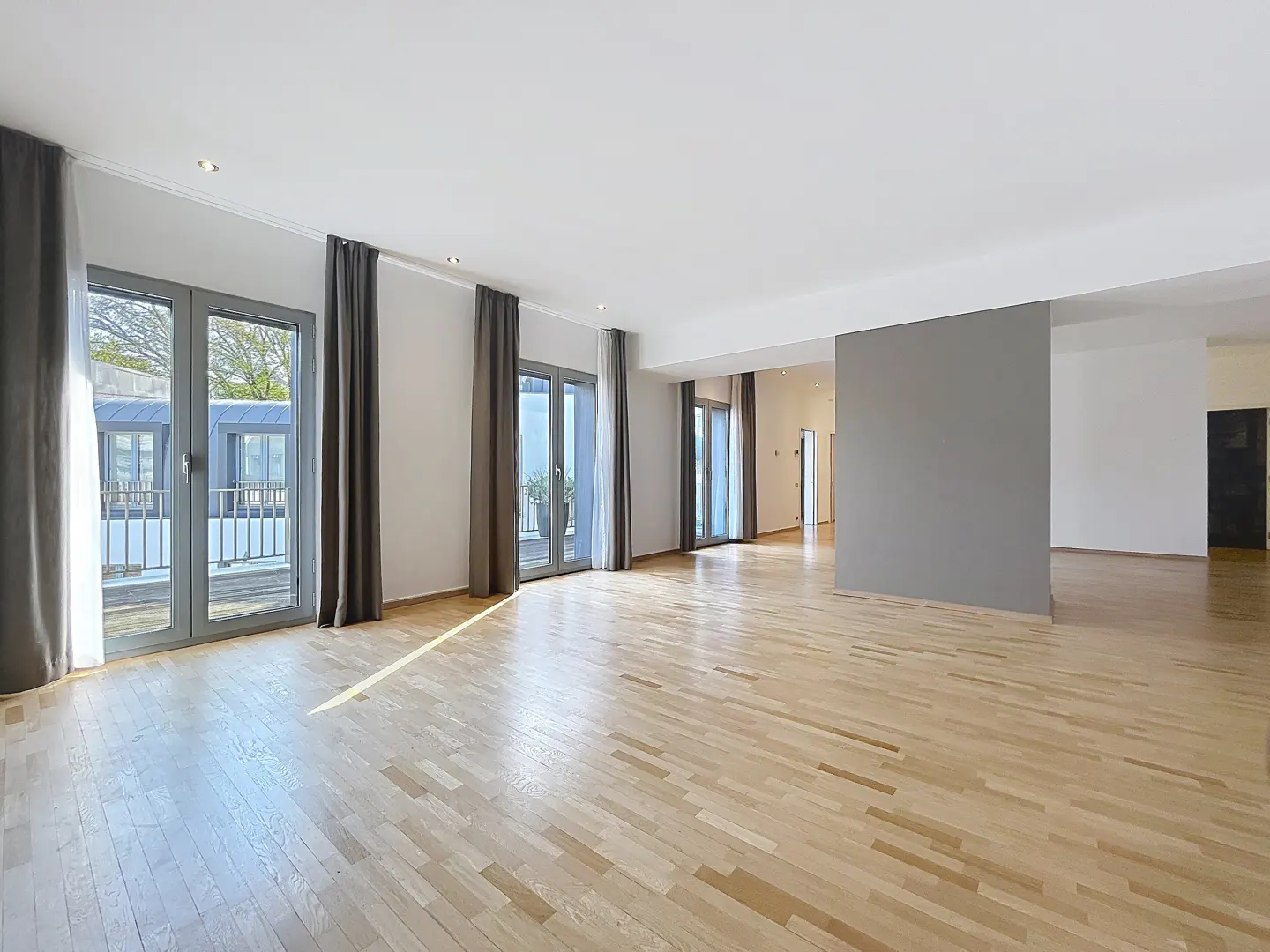 Bright, empty room with light wood floors, white walls, and gray accents. Balcony access through gray-framed glass doors with gray curtains.
