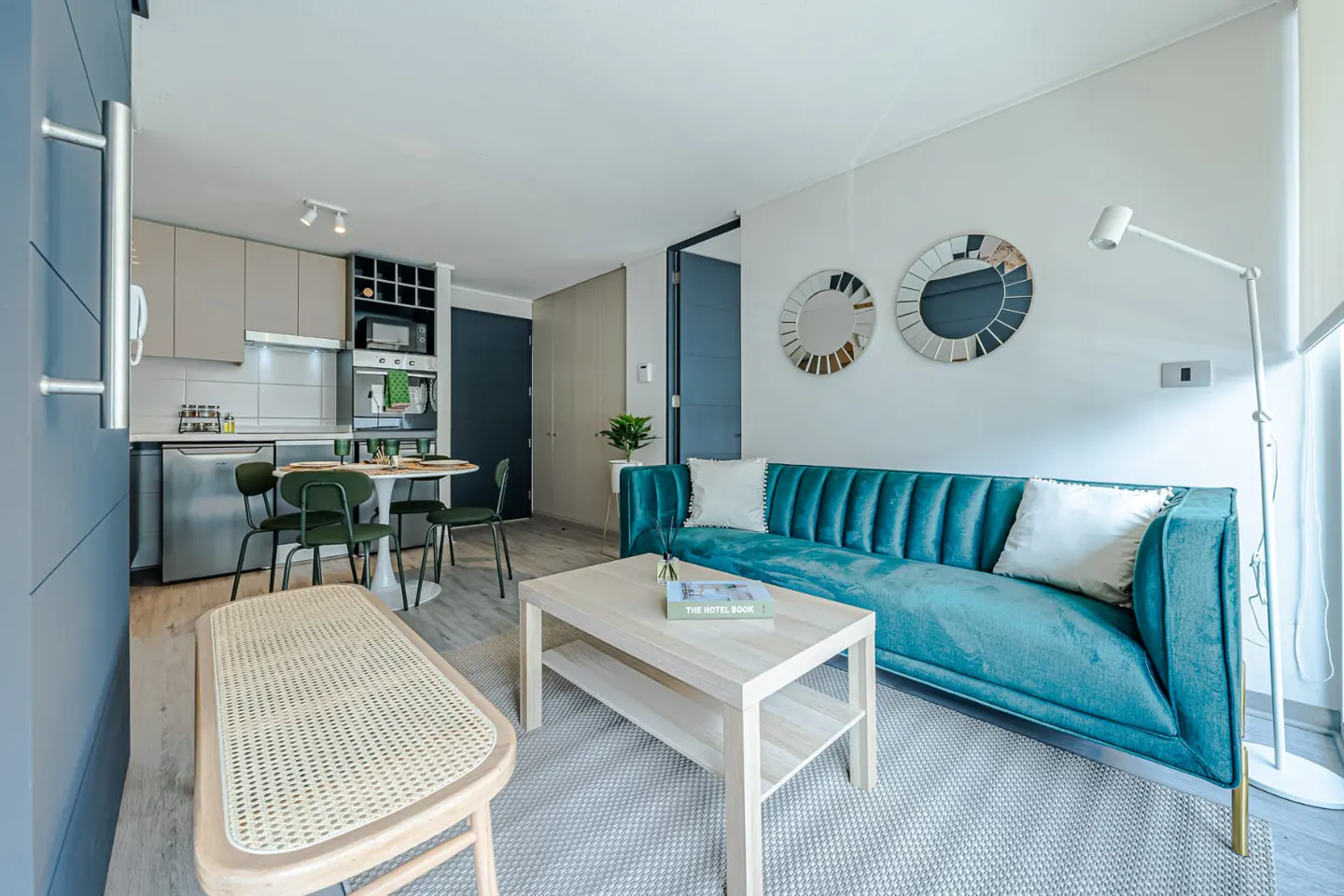 Modern open-plan apartment with a teal velvet sofa, light wood coffee table, and a small dining area with a kitchenette.