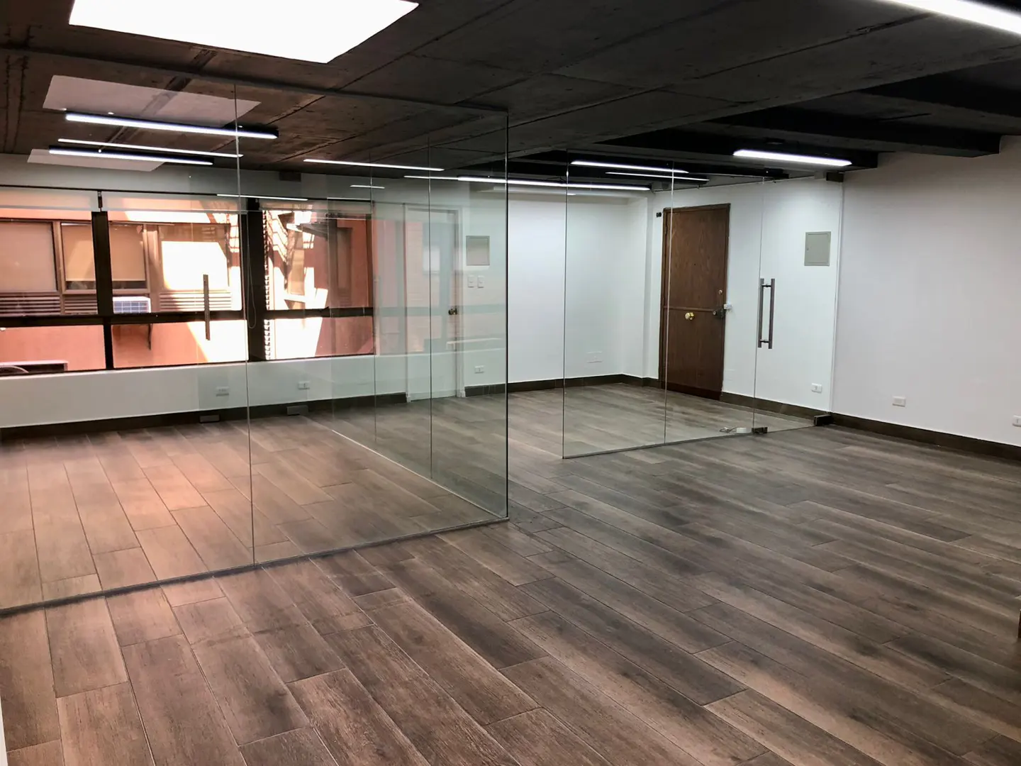 Empty office space with wood-look tile floors, glass walls, white walls, and a dark ceiling. A window is on the left.