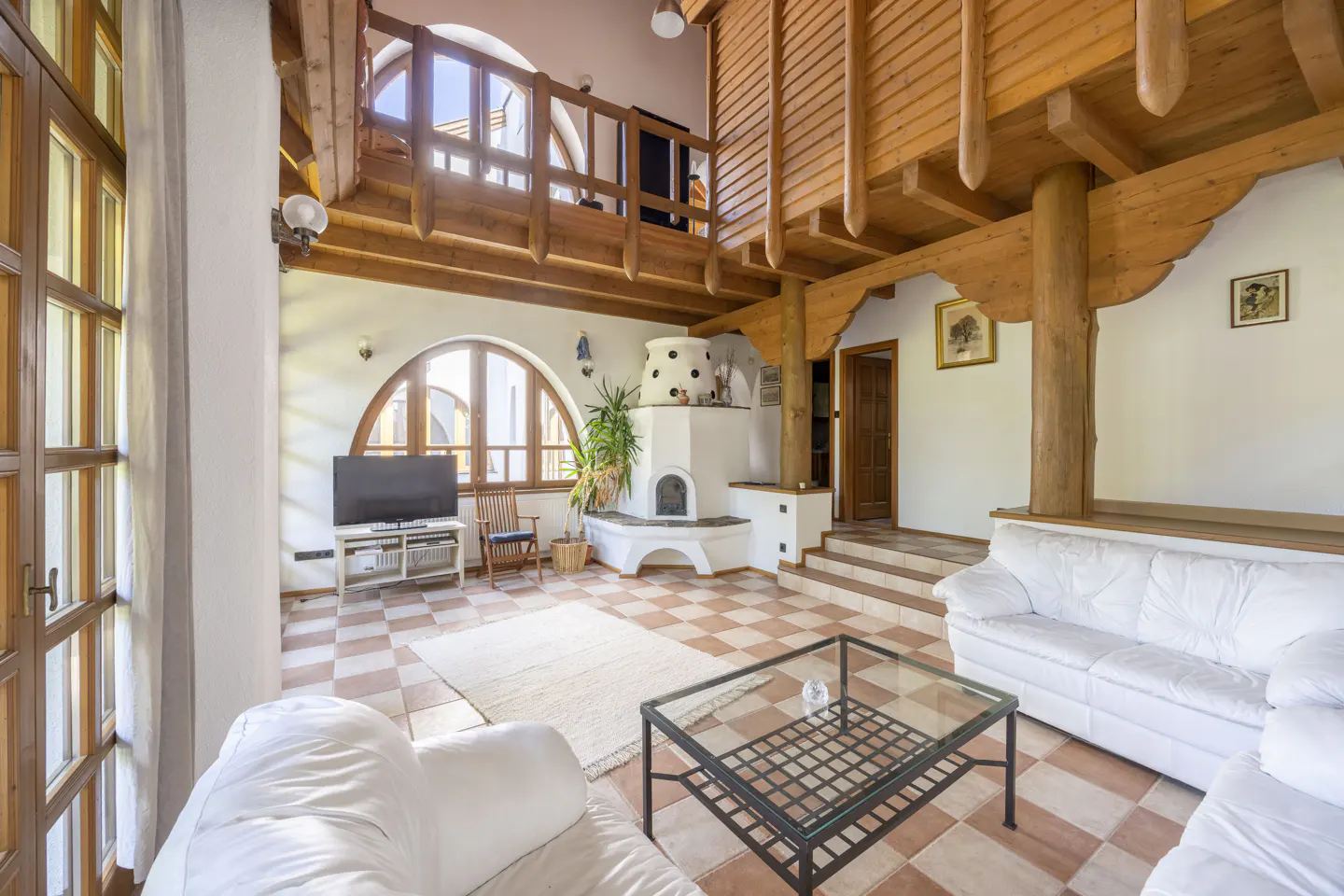 Bright living room with white sofas, a glass table, and a white fireplace. Arched windows and wooden beams add character.
