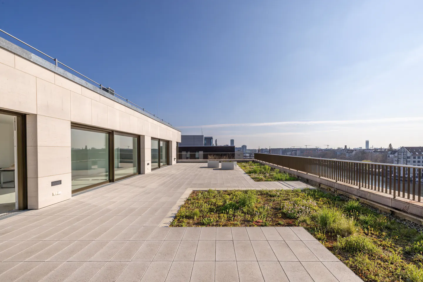 Rooftop terrace with stone tiles, green vegetation, and a metal railing overlooking a city skyline under a clear blue sky.