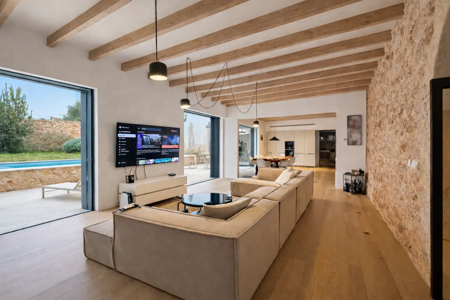Bright living room with wood floors, exposed beams, and stone wall. A large beige sectional faces a TV and open patio doors to a pool.