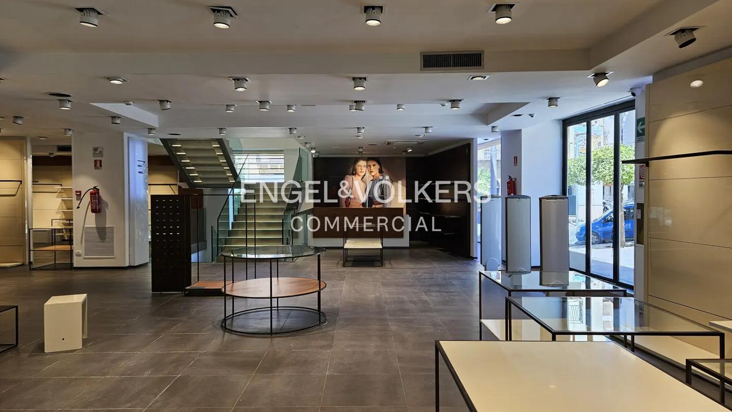 Interior of a commercial space with display tables, stairs, and an Engel & Völkers sign. The floor is gray tile, and there are many ceiling lights.