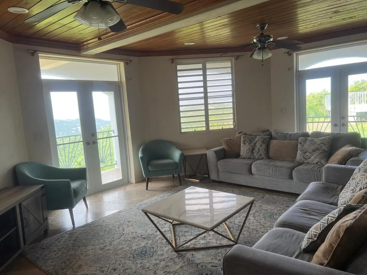 Living room with a gray sectional sofa, two teal chairs, and a marble coffee table on a patterned rug. Balcony doors offer a view of lush greenery.