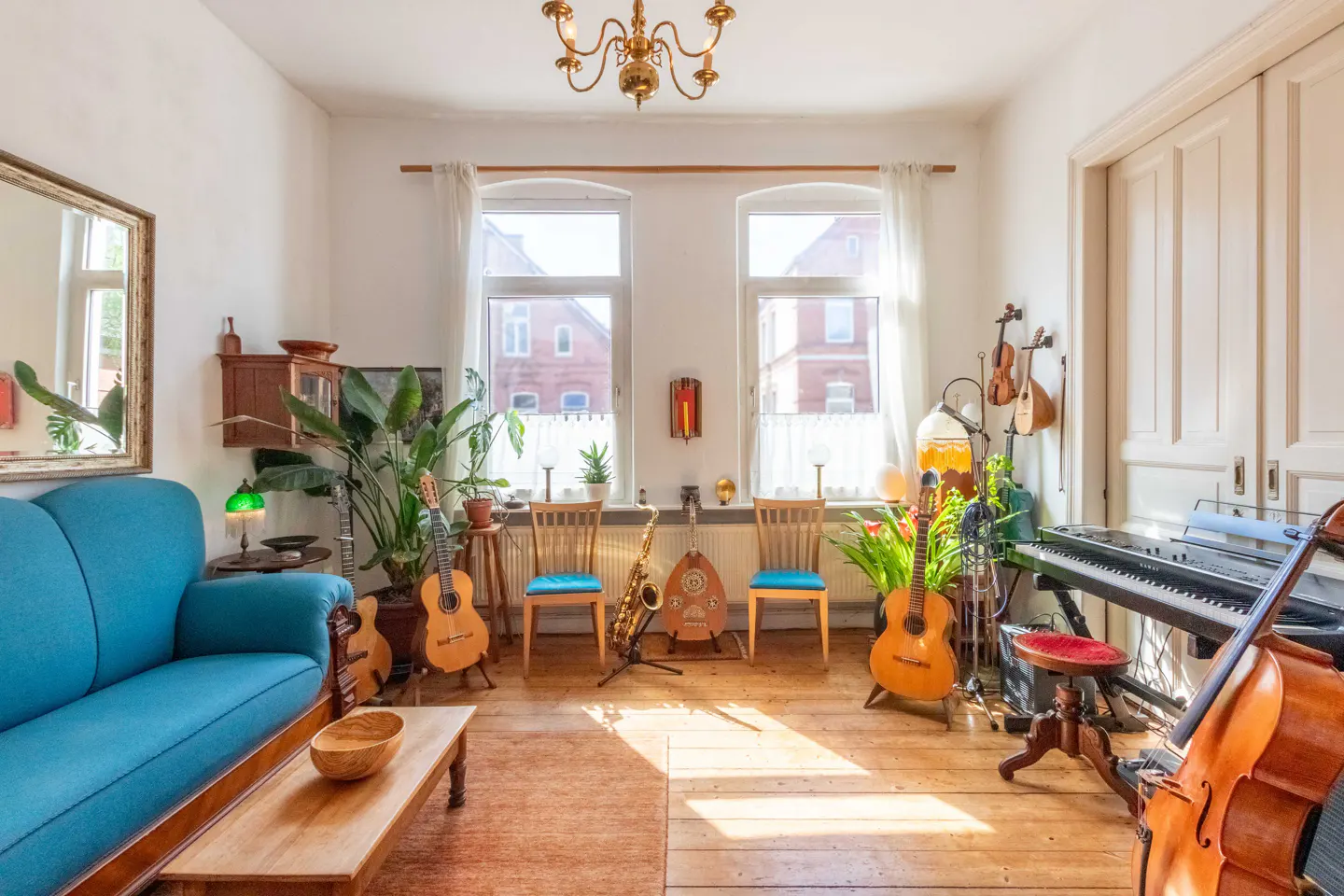 Bright living room with a blue sofa, wooden floors, two windows, and various musical instruments including guitars and a piano.