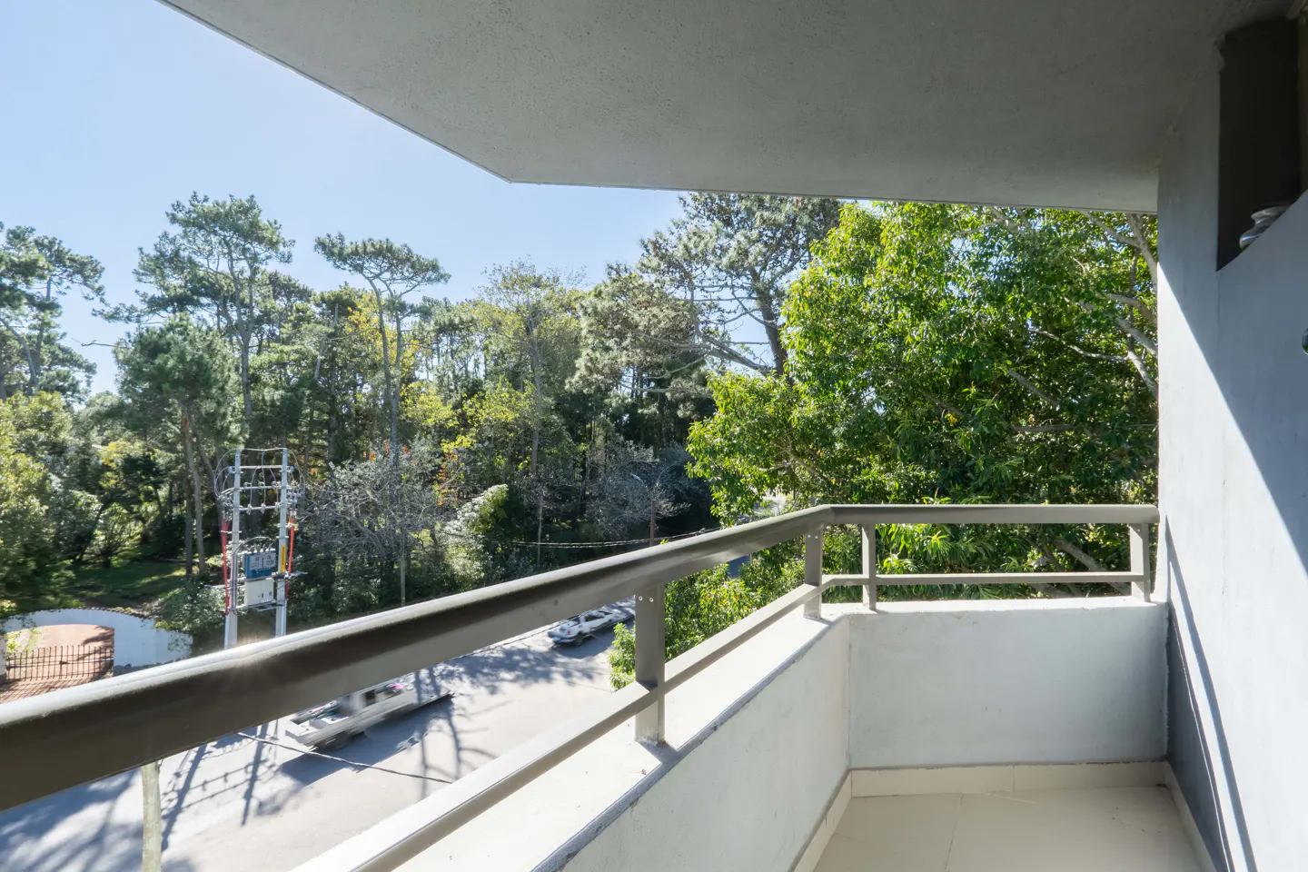 View from a gray balcony overlooking a street with trees and cars on a sunny day.