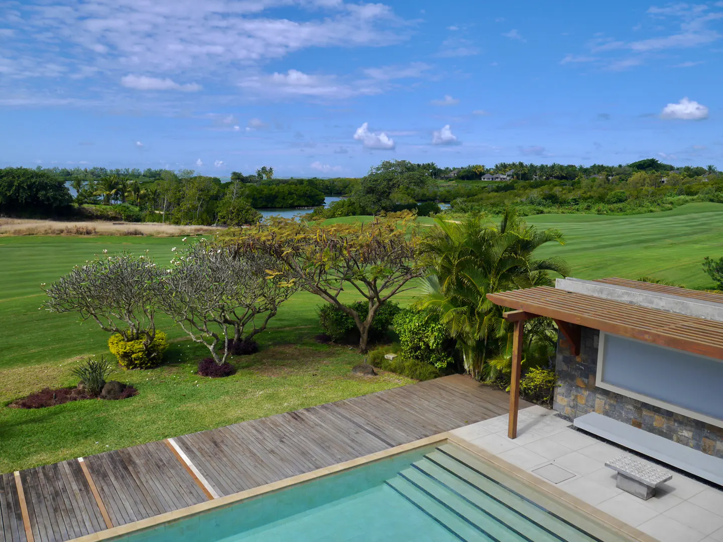 A swimming pool with a wooden deck overlooks a lush green golf course, tropical trees, and a blue sky with light clouds.